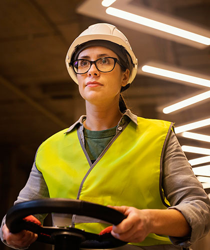 Operaria con casco de seguridad, gafas y chaleco reflectante sujeta el volante de un vehículo industrial